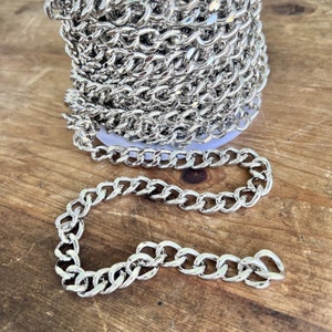May include: A silver curb chain with a shiny finish. The chain is coiled on a spool and a section of the chain is laid out on a wooden surface.