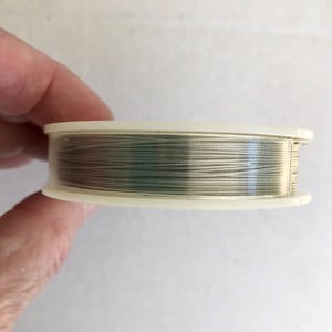 May include: A spool of silver-colored thread wound around a clear plastic bobbin. The thread is thin and appears to be metallic.