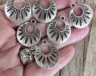 May include: A collection of silver-toned, circular pendants with a sunburst design. Each pendant has a central hole and a small loop at the top for attaching to jewelry. The pendants are displayed on a wooden surface.