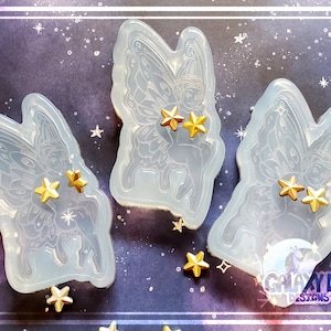 May include: Three clear silicone molds of fairies with wings, each mold has two gold stars on the fairy's wings. The molds are on a purple background with gold stars.
