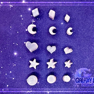 May include: A collection of 15 white resin charms in various shapes, including stars, hearts, moons, and diamonds. The charms are arranged on a purple background with glitter.