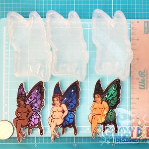 May include: Two silicone molds for making resin crafts. Each mold is shaped like a fairy with butterfly wings. The fairies are sitting on a stool with their legs crossed. One fairy has purple wings and the other has blue wings.