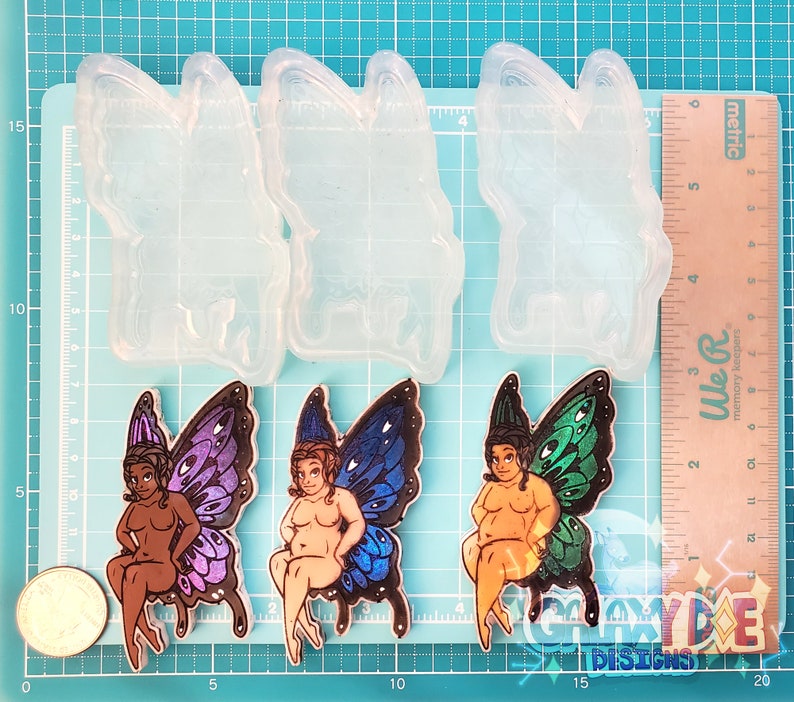 May include: Two silicone molds for making resin crafts. Each mold is shaped like a fairy with butterfly wings. The fairies are sitting on a stool with their legs crossed. One fairy has purple wings and the other has blue wings.