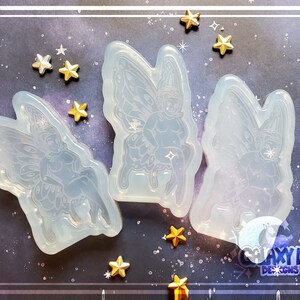 May include: Three clear silicone molds of fairies with wings, sitting on a mushroom. The molds are on a dark blue background with gold stars.