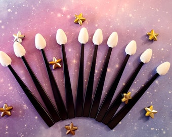 May include: Ten black makeup sponge applicators with white foam tips arranged in a semi-circle on a purple and pink background with gold stars.