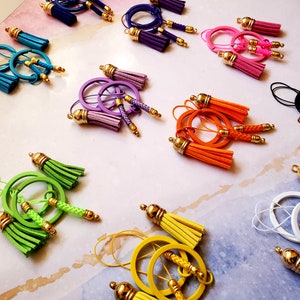 May include: Assortment of colorful tassel earrings with gold hoops and metal accents. The earrings feature a variety of colors, including green, orange, yellow, purple, blue, pink, and black.