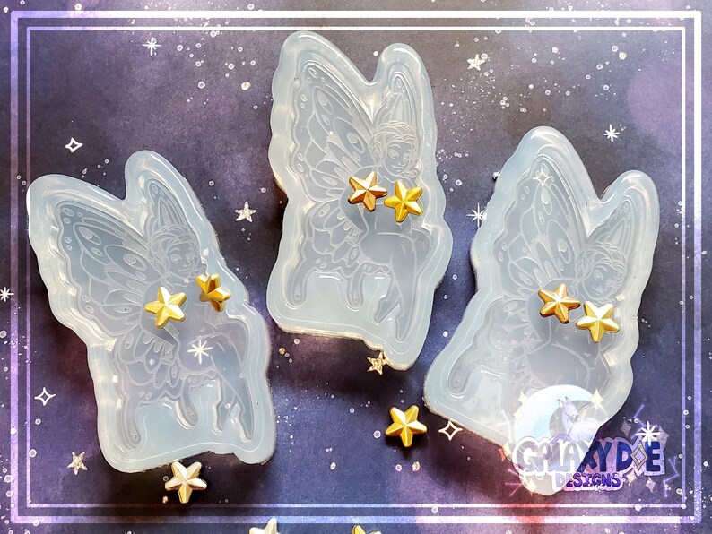 May include: Three clear silicone molds of fairies with wings, each mold has two gold stars on the fairy's wings. The molds are on a purple background with gold stars.
