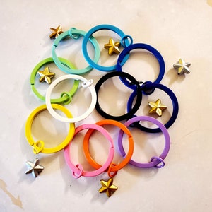 May include: A set of ten colorful metal key rings with gold star charms. The key rings are arranged in a circle with the stars facing outwards.