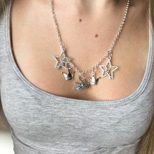 May include: Silver-toned necklace featuring star and crown charms. The necklace has a delicate chain and multiple charms, including openwork stars and small crown pendants. The jewelry is worn over a gray tank top.