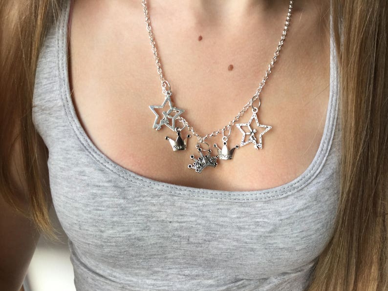 May include: Silver-toned necklace featuring star and crown charms. The necklace has a delicate chain and multiple charms, including openwork stars and small crown pendants. The jewelry is worn over a gray tank top.