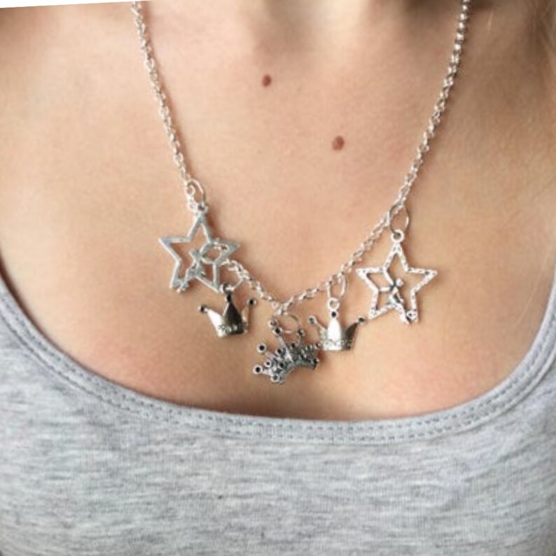 May include: Silver-toned necklace featuring star and crown charms. The necklace has a delicate chain and multiple star pendants, along with small crown charms. The jewelry is worn against a gray top.