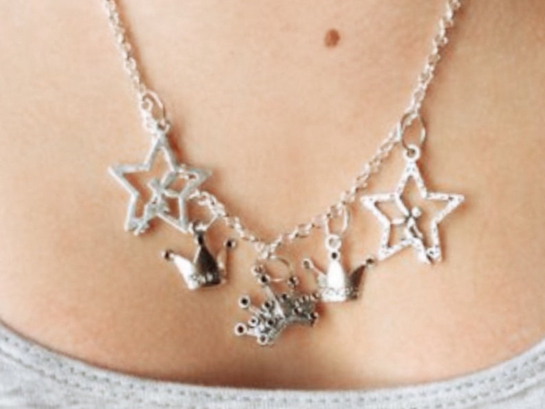 May include: Silver-toned necklace featuring star and crown charms. The necklace has a delicate chain and multiple charms, including openwork stars and small crown pendants. The jewelry is worn around the neck.