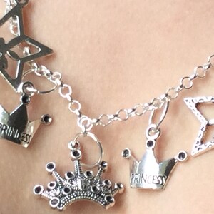 May include: Silver-toned charm bracelet featuring star and crown pendants. The crowns have the word "PRINCESS" engraved on them. The bracelet is designed with a delicate chain and is suitable for a variety of occasions.