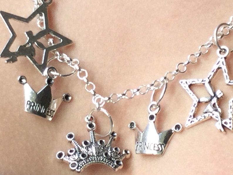 May include: Silver-toned charm bracelet featuring star and crown pendants. The crowns have the word "PRINCESS" engraved on them. The bracelet is designed with a delicate chain and is suitable for a variety of occasions.
