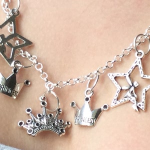 May include: Silver-toned necklace with multiple charms. The charms include open stars and crowns, some with the word "PRINCESS" inscribed. The necklace is made of linked chain and is worn around the neck.
