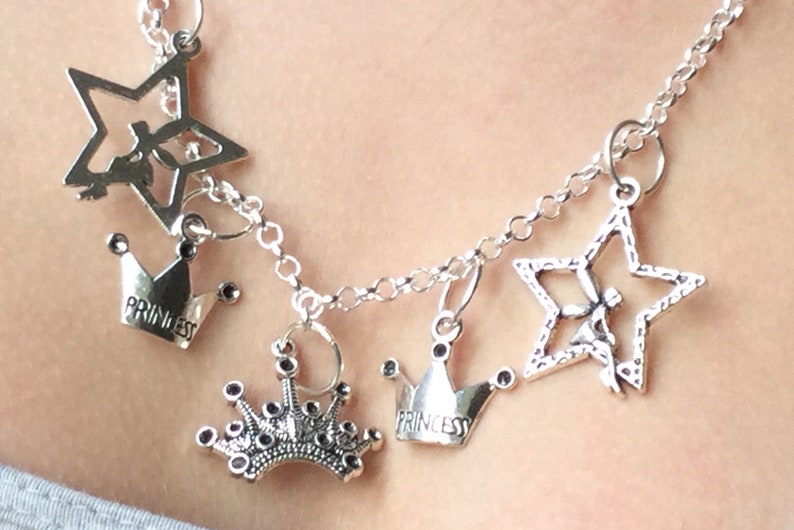 May include: Silver-toned necklace with multiple charms. The charms include open stars and crowns, some with the word "PRINCESS" inscribed. The necklace is made of linked chain and is worn around the neck.