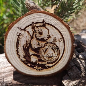 May include: A wooden circle with a squirrel design and a black and white circle with the text "Unity, Service, Recovery" and "XVI" inside. The text "Lori" is written on the top right of the circle. The text "10-4-2003" is written below the circle.