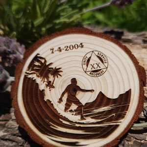 Op de afbeelding: Een houten cirkel met een surfer-silhouet, palmbomen en de tekst "7-4-2004". Een driehoek met de woorden "Eenheid", "Dienst" en "Herstel" is in de cirkel gegraveerd. De cirkel heeft een bruine houtnerf.