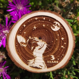 Op de afbeelding: Een houten cirkel met een kattenkop en een halve maan met sterren op de achtergrond. De tekst "3 JAAR" is gegraveerd onder de kattenkop.