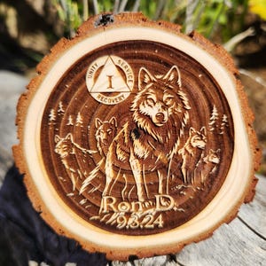 May include: A round wooden plaque with a carved wolf pack design. The plaque features a central wolf surrounded by others, with trees and the text "Ron D. 9.8.24". A smaller emblem reads "Unity, Service, Recovery".
