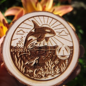 May include: A round wooden coaster with a detailed laser-etched design. The design features an orca breaching the water, a turtle, fish, coral, and the name "Candice". Includes the date "6-1-2024" and the words "Unity, Service, Recovery".
