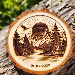 May include: A wooden round slice with a laser-engraved design of a mountain landscape with a sun, a river, and trees. The text "2 Years" and the date "01-01-2023" are also engraved on the slice.