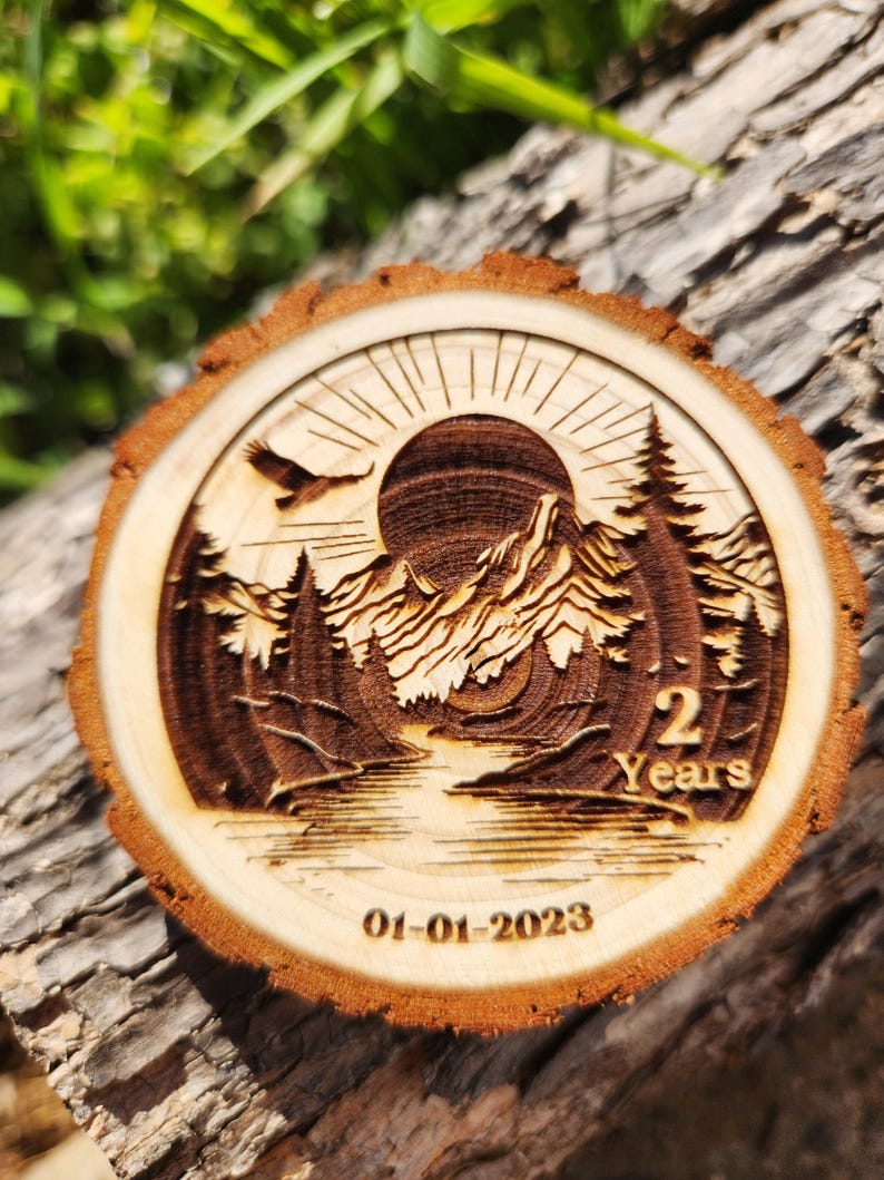 May include: A wooden round slice with a laser-engraved design of a mountain landscape with a sun, a river, and trees. The text "2 Years" and the date "01-01-2023" are also engraved on the slice.