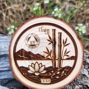 May include: A circular wooden coaster featuring a laser-etched design. The design includes the name "Elaine", bamboo, a lotus flower, mountains, and the text "Unity, Service, Recovery". The coaster has a natural wood grain edge.
