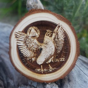 May include: A round wooden plaque with a detailed wood-burned image of a chicken with outstretched wings. The plaque includes the text "UNITY SERVICE RECOVERY" and the date "1/31/22". The wood has a natural, light brown color.