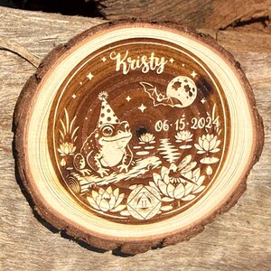 May include: A circular wood slice with a laser-engraved design featuring a frog wearing a party hat, the name Kristy, the date 06-15-2024, a bat flying past a moon, water lilies, and a small geometric emblem with the Roman numeral II at the bottom.