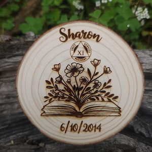 Può includere: Targa rotonda in legno con il nome "Sharon" inciso in corsivo. Presenta un disegno floreale sopra un libro aperto, con la data "6/10/2014" sotto. La targa riporta anche la scritta "Unity, Service, Recovery" e il numero romano "XI".