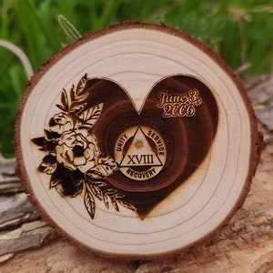 May include: A wooden slice with a heart-shaped design. The heart contains the text "June 3, 2006" and a symbol with the words "Unity, Service, Recovery" and the Roman numeral "XVIII". Floral elements are etched around the heart.