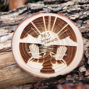 May include: A wooden round disc with a sunburst design and the text "365 Sober Sunsets" engraved on it. Two chairs are depicted below the sunburst, facing each other.