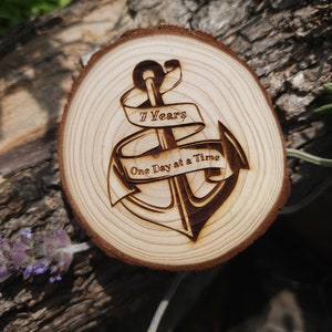Può includere: Una fetta rotonda di legno con un design bruciato di un'ancora con uno striscione che recita "7 Years One Day at a Time".