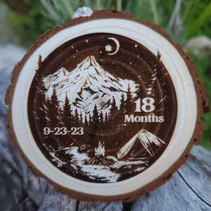 May include: A round wooden slice with a detailed camping scene etched into the surface. The design includes a mountain range, trees, a tent, a campfire, and the text "18 Months" and "9-23-23". The outer edge of the wood is a darker brown.