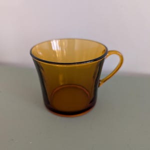 K&ouml;nnte beinhalten: Bernsteinfarbene Teetasse aus Glas mit rundem Henkel und schwarzem Rand. Die Tasse hat eine leicht ausgestellte Form und eine glatte, gl&auml;nzende Oberfl&auml;che. Die Teetasse ist leer und steht auf einer hellgr&uuml;nen Oberfl&auml;che.