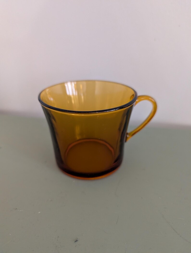 K&ouml;nnte beinhalten: Bernsteinfarbene Teetasse aus Glas mit rundem Henkel und schwarzem Rand. Die Tasse hat eine leicht ausgestellte Form und eine glatte, gl&auml;nzende Oberfl&auml;che. Die Teetasse ist leer und steht auf einer hellgr&uuml;nen Oberfl&auml;che.