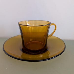 K&ouml;nnte beinhalten: Bernsteinfarbenes Teetassen- und Untertassen-Set aus Glas. Die Tasse hat einen gebogenen Henkel und steht auf einer passenden Untertasse. Das Set hat einen dunklen Rand um die Untertasse. Das Glas hat eine Vintage-&Auml;sthetik.