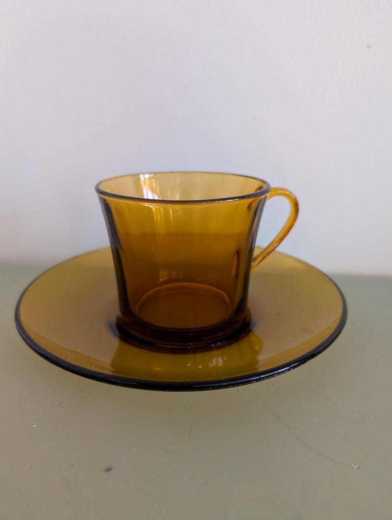 K&ouml;nnte beinhalten: Bernsteinfarbenes Teetassen- und Untertassen-Set aus Glas. Die Tasse hat einen gebogenen Henkel und steht auf einer passenden Untertasse. Das Set hat einen dunklen Rand um die Untertasse. Das Glas hat eine Vintage-&Auml;sthetik.