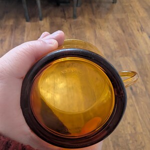 K&ouml;nnte beinhalten: Bernsteinfarbener Glasbecher mit dunkelbraunem Rand und Henkel. Auf dem Boden des Bechers steht das Wort "DURALEX" in umgekehrter Reihenfolge. Der Becher besteht aus transparentem Glas.