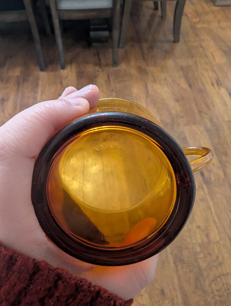 K&ouml;nnte beinhalten: Bernsteinfarbener Glasbecher mit dunkelbraunem Rand und Henkel. Auf dem Boden des Bechers steht das Wort "DURALEX" in umgekehrter Reihenfolge. Der Becher besteht aus transparentem Glas.