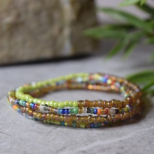 May include: Three beaded bracelets in shades of green, gold, and various colours. The bracelets are layered, creating a stacked effect. The small, round beads give a delicate, bohemian look. The bracelets are suitable for everyday wear.