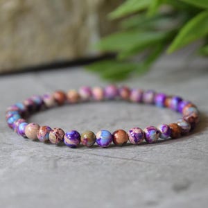 Peut inclure: Un bracelet de perles rondes de différentes couleurs, notamment violet, bleu et marron. Le bracelet est posé sur une surface grise. Un bijou.