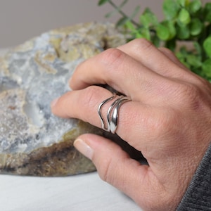 May include: A silver ring with a unique, flowing design. The ring features three curved bands that create a wave-like appearance. The jewelry is displayed on a hand, showcasing its elegant and modern style.