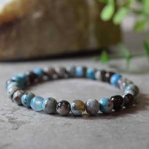 May include: A stretch bracelet composed of round beads in varying shades of blue, grey, and dark brown. The beads feature a polished, smooth finish with marbled patterns and translucent sections, arranged in a repeating sequence on an elastic cord.
