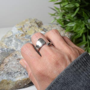 Peut inclure: Une bague argentée avec un dessus incurvé et plat est portée au doigt. La bague a un design ouvert. L'arrière-plan comprend une grande roche texturée et un feuillage vert.