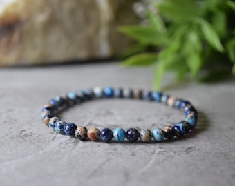 May include: A beaded bracelet featuring a variety of blue, brown, and cream-colored jasper stones. The bracelet is strung on an elastic band and is displayed on a gray surface. The background includes a blurred rock and green foliage.