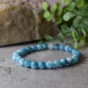 May include: A bracelet composed of light blue, spherical beads. The beads have a smooth, polished finish and are strung together on an elastic band. The bracelet is displayed on a grey surface with a blurred background of a rock and green foliage.
