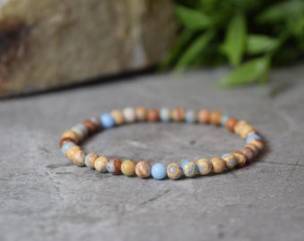 May include: A beaded bracelet featuring small, round, polished stones in a variety of earthy tones including tan, brown, and beige, interspersed with smooth, solid light blue beads arranged in a circular pattern on a flexible cord.