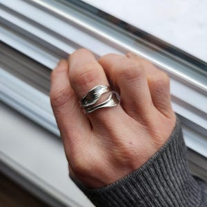 May include: A silver ring with a flowing, organic design. The ring has a layered, wave-like pattern, creating a textured and sculptural appearance. The ring is worn on a finger, showcasing its intricate details and reflective surface.